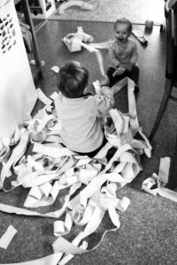 Dvě malé děti sedící na podlaze se smějí a vymotávají pokladní pásku