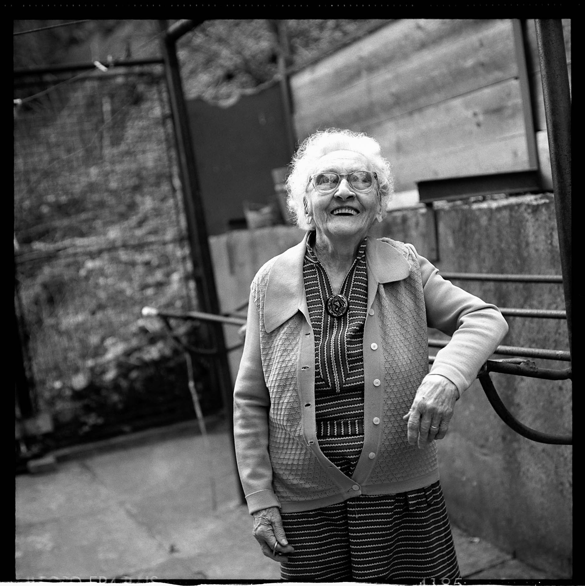 Portrét smějící se staré paní v pruhovaných šatech opírající se o klepač na dvorku