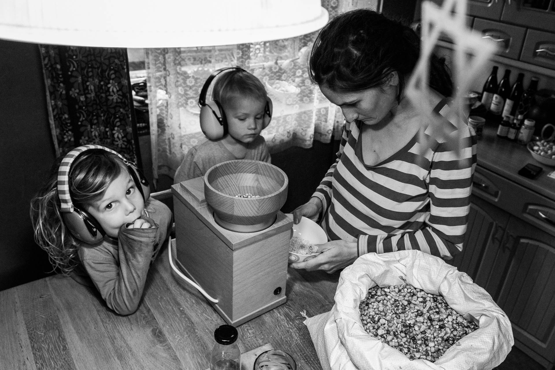 Dvě děti se sluchátky na uších pozorují svoji mámu jak mele kukuřičnou mouku